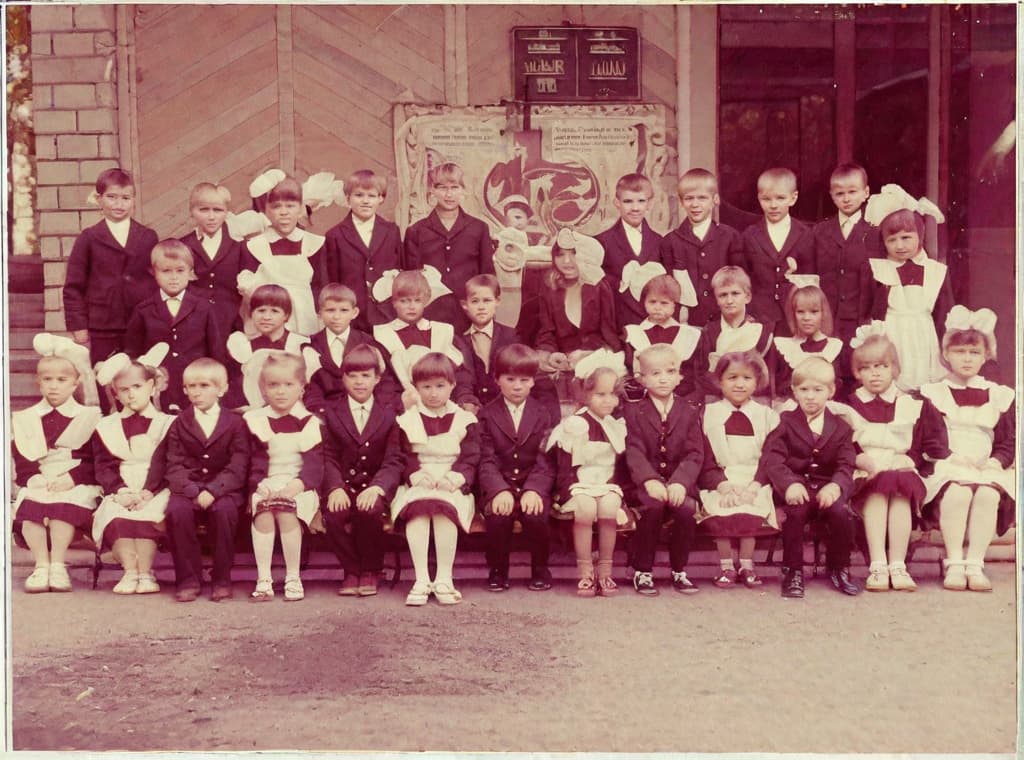  It was taken on September 1st 1986, exactly 128 days or 40 months and 4 days after the Chernobyl catastrophe took place. first day at in Gomel, Belarus. I had just turned seven year that summer. In the photo one can see my clmates. mates and our teacher. Some are standing with their arms hanging besides their bodies, while others are seated on a low bench with their hands neatly placed. placed on their . The students are arranged in three lines. Ten in the third top line, nine in the second middle line. the line and 13 in the first front line, totaling 32 students. There are 17 boys and 15 s. distinguished by the gr coat. My friend Gennady is the fourth from the right in the third top line. I am seated in the very