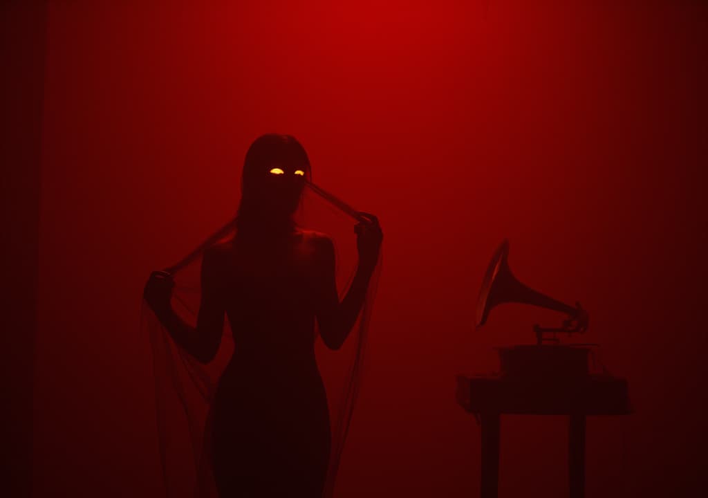  good quality, high quality, dimly lit, deep red room. woman moves languidly, veiled in sheer fabric. her dance reveals and conceals, seducing. luminous, enigmatic eyes hold dangerous allure. haunting melody from old phonograph. hypnotic, sensory scene of mystery and desire.