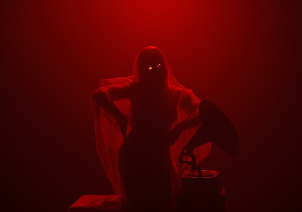  good quality, high quality, dimly lit, deep red room. woman moves languidly, veiled in sheer fabric. her dance reveals and conceals, seducing. luminous, enigmatic eyes hold dangerous allure. haunting melody from old phonograph. hypnotic, sensory scene of mystery and desire.