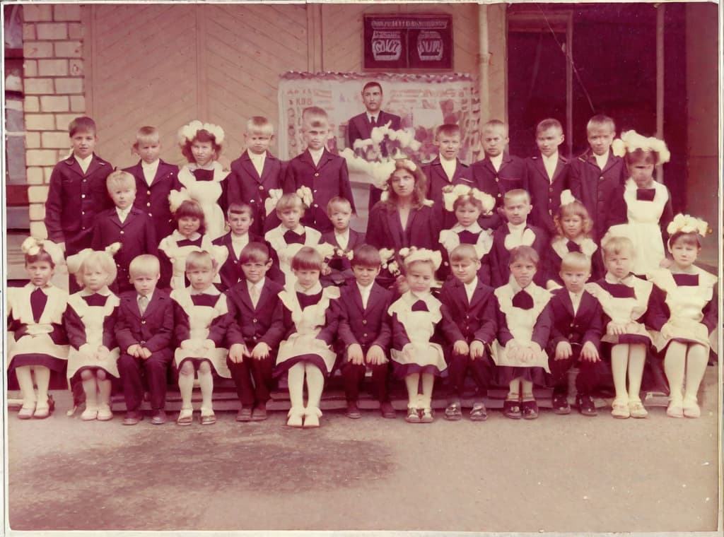  nine hundred and twenty eight days or four months and four days after the Chernobyl catastrophe took place. It captures my first day in Gomel, Belarus. I had just turned seven year that summer. In this photo, one can see my clmates. and our teacher. Some are standing with their arms hanging besides their bodies while others are seated on the on on the low bench with with our hands neatly placed on their . The students are arranged in three lines, ten in the third top back line, three. 9 in the second middle line and 13 in the first front line, totally in 13. There are 17 boys and 15 s distinguished by the dress code. My friend Gennady is the fourth from the right on the third top line.;