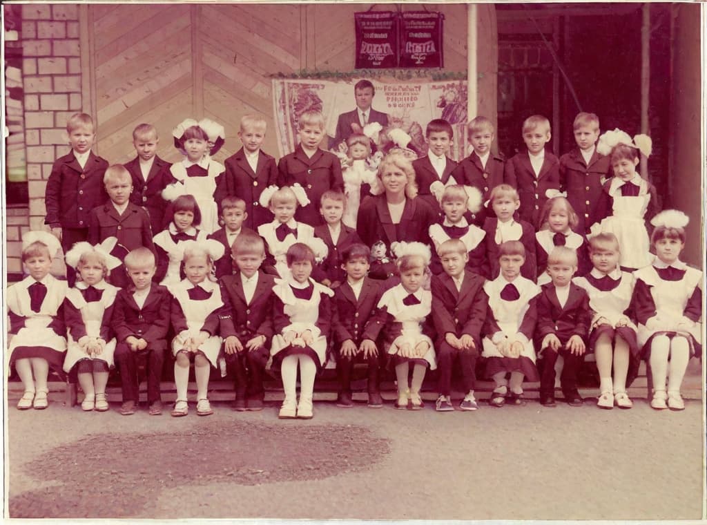  It was taken on September 1st 1986, exactly 128 days or 40 months and 4 days after the Chernobyl catastrophe took place. first day at in Gomel, Belarus. I had just turned seven year that summer. In the photo one can see my clmates. mates and our teacher. Some are standing with their arms hanging besides their bodies, while others are seated on a low bench with their hands neatly placed. placed on their . The students are arranged in three lines. Ten in the third top line, nine in the second middle line. the line and 13 in the first front line, totaling 32 students. There are 17 boys and 15 s. distinguished by the gr coat. My friend Gennady is the fourth from the right in the third top line. I am seated in the very