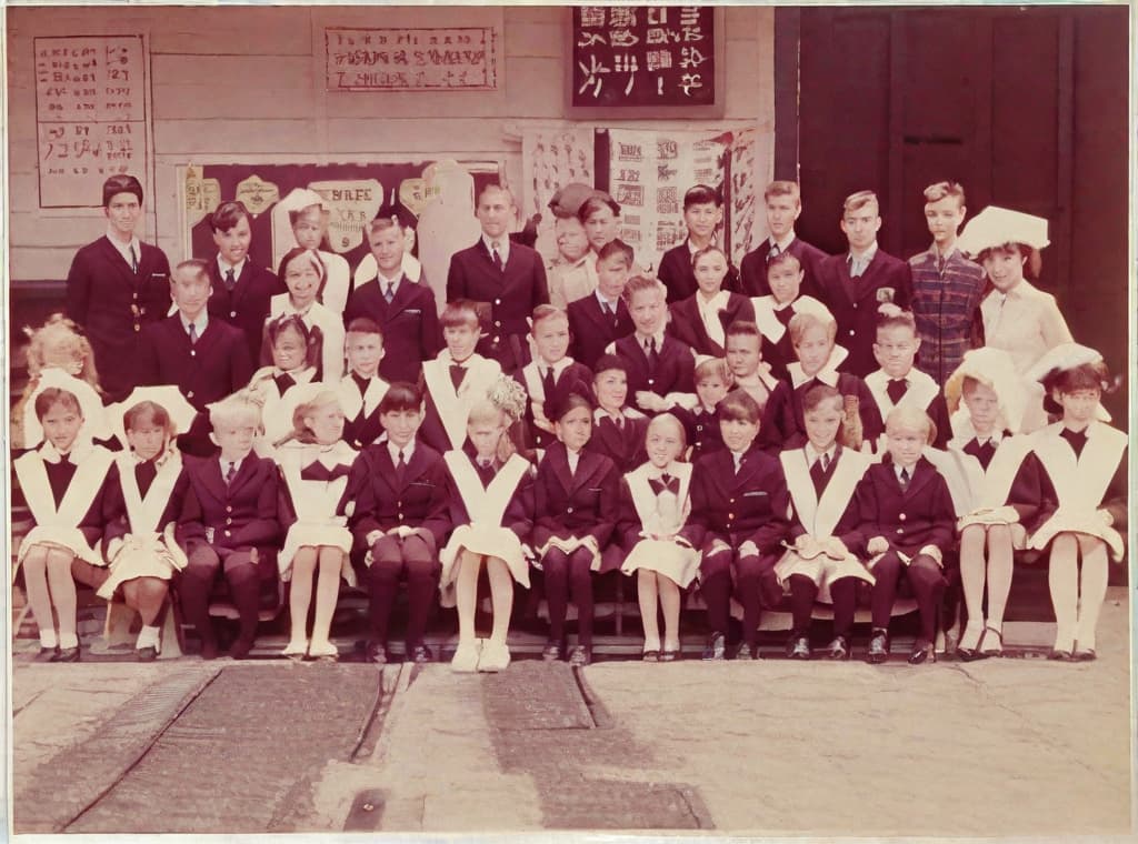  Standing back row with ren in uniform, two s (one male in a suit and tie, one female with hair pulled back and wearing a light and dark ), against a wooden building background with educational posters next to the entrance