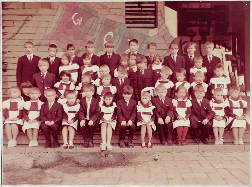  This photo was taken on September 1st 1986, exactly 128 days, or 4 months and 4 days, after the Chernobyl catastrophe took place. It captures my first day at in Gomel, Belarus. I had just turned that summer. In the photo, one can see my clmates and our teacher. Some are standing with their arms hanging beside their bodies, while others are seated on a low bench with their hands neatly placed on their . The students are arranged in three lines: 10 in the 3rd (top) back line, 9 in the 2nd (middle) line, and 13 in the 1st front line, totaling 32 students. There are 17 boys and 15 s, distinguished by the dress code. My friend Genadi is 4th from the right in the 3rd (top) line. I am seated in the very center of the