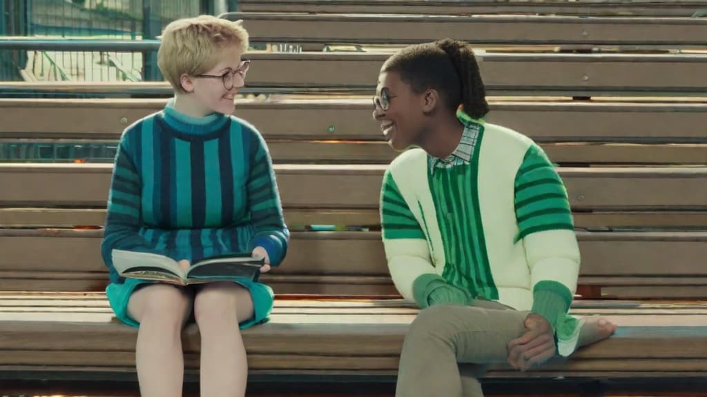  The image features two individuals, likely in a casual, friendly or sentimental setting, sitting on a wooden bench that shows signs of wear and weathering. The environment suggests an outdoor public space, possibly a park. The person on the left is a short haired individual wearing glasses, a teal blue sweater, and blue shorts. Their posture is relaxed, sitting with one leg crossed over the other, and they're smiling broadly while looking at the other person, indicating a moment of joyful interaction. The person on the right wears a vertically striped green and white shirt with rolled up sleeves and black pants. They are slightly turned towards the person on the left, with a subdued smile and a contemplative expression, suggesting engagem