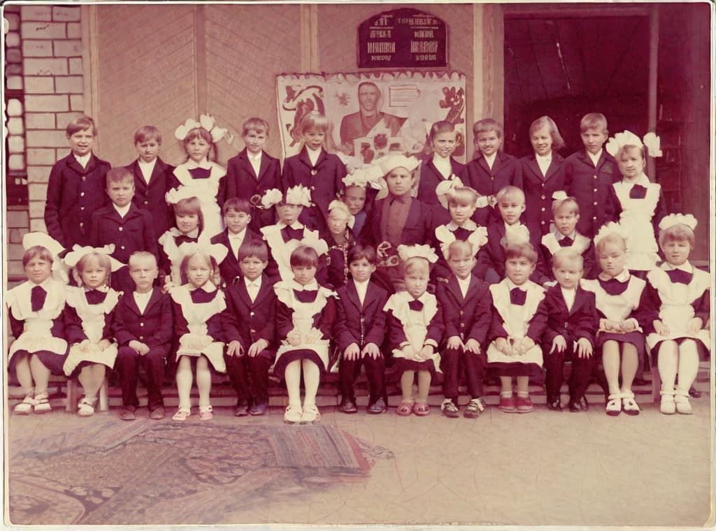  It was taken on September 1st 1986, exactly 128 days or 40 months and 4 days after the Chernobyl catastrophe took place. first day at in Gomel, Belarus. I had just turned seven year that summer. In the photo one can see my clmates. mates and our teacher. Some are standing with their arms hanging besides their bodies, while others are seated on a low bench with their hands neatly placed. placed on their . The students are arranged in three lines. Ten in the third top line, nine in the second middle line. the line and 13 in the first front line, totaling 32 students. There are 17 boys and 15 s. distinguished by the gr coat. My friend Gennady is the fourth from the right in the third top line. I am seated in the very